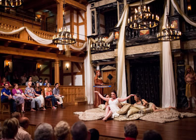 The wood, two-level playhouse draped in white fabric and gold netting, chandeliers hanging over the stage, and on stage Cleopatra in white night gown with her arms outstretch as she sits at the front of the floor cushions, lounging behind her are Iras and Charmian, and standing at the back corners of the stage are Mardian in gold robes and waving a fan, and bare-torsoed Alexes in Egyptian headress and skirt, plus audience sitting on gallant stoos on the stage as well as in seats to the side and in front of the stage.